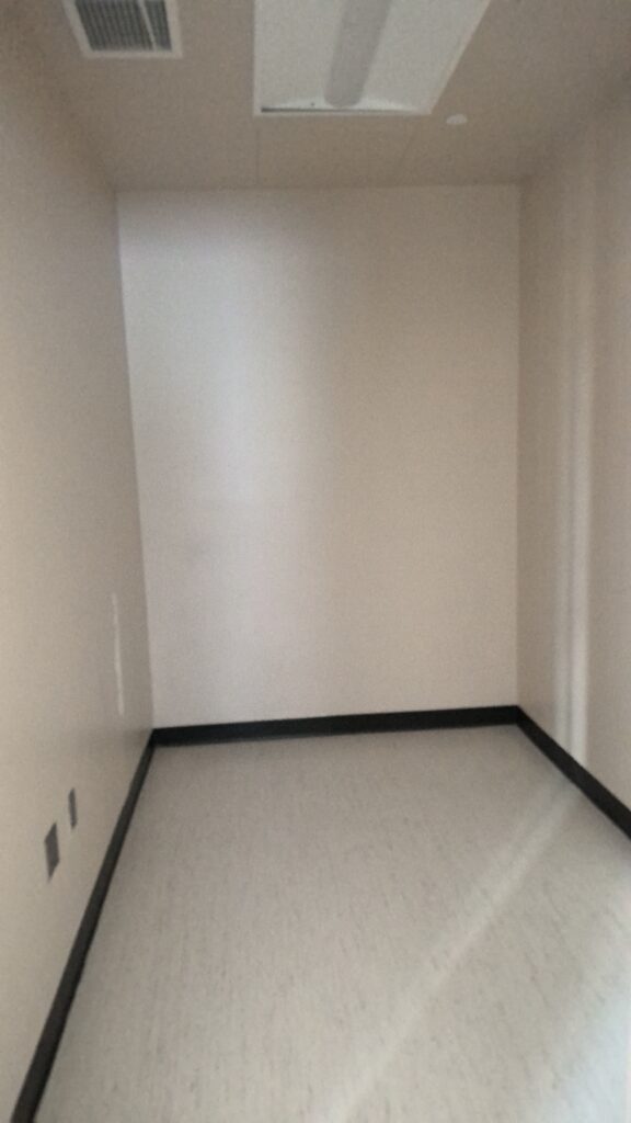 A small room with white walls and white flooring. A recessed rectangular fluorescent light and a vent are on the ceiling.