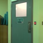 A green metal door with a square window and a printed sign that says, "take a break, take a breath!" Next to the door is a round button glowing red.