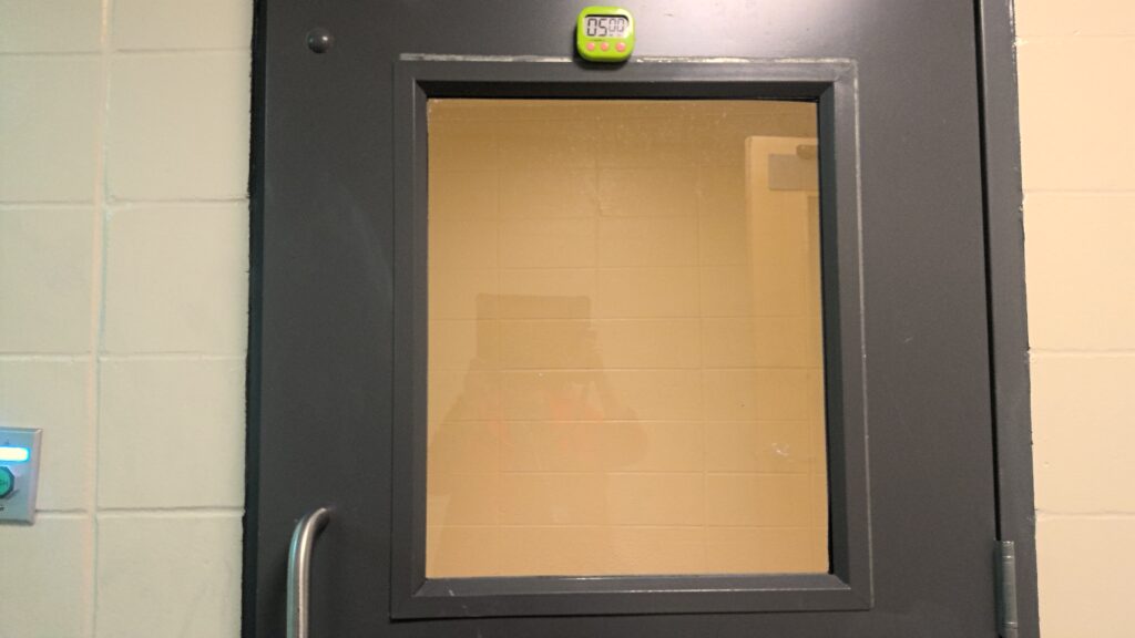 Metal door with large observation window. Green timer affixed to the top of the door reads 05:00.