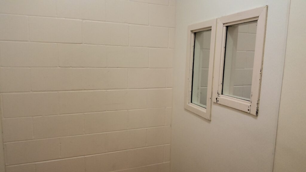 Inside corner of a room with white cinder block wall and two small windows. The white paint around the window frames is chipping off.