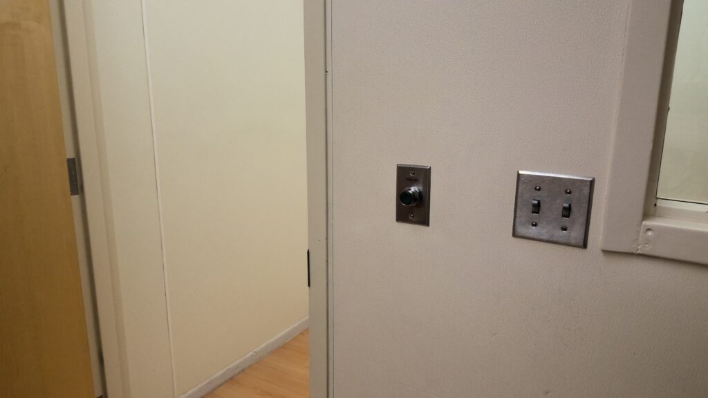 Photograph of an indoor corner showing a beige wall with two light switches and a thermostat. A wooden door is partially visible on the left, open.