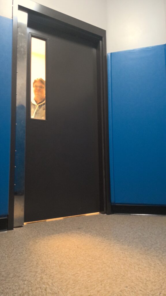 A room with blue padded wall panels and a gray metal door with a narrow window along the upper edge. An adult is visible through the window standing on the opposite side of the door.