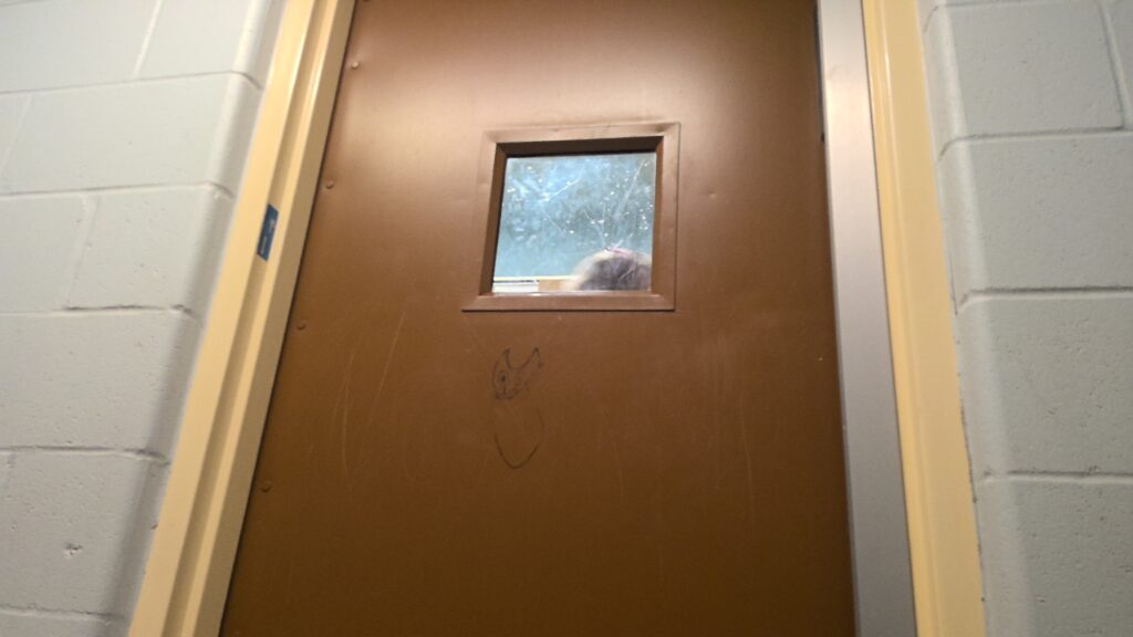 A brown metal door with a small square window. There are scratches and graffiti on the door and a dirty observation window.