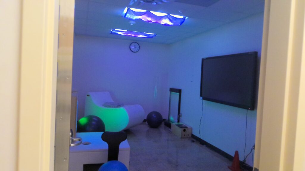A room with a rocking sensory chair and a TV mounted to the wall. Blue fabric over the ceiling light casts the room in low blue light.