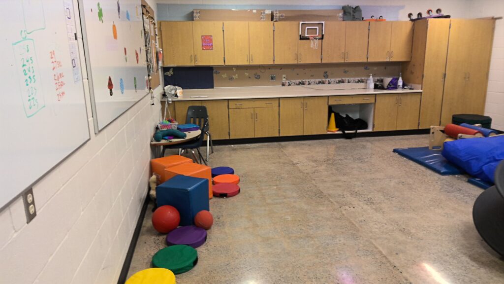 A large room with counters and cabinets against one wall and whiteboards against another. There are multicolored cushions, weighted balls, and gym mats.