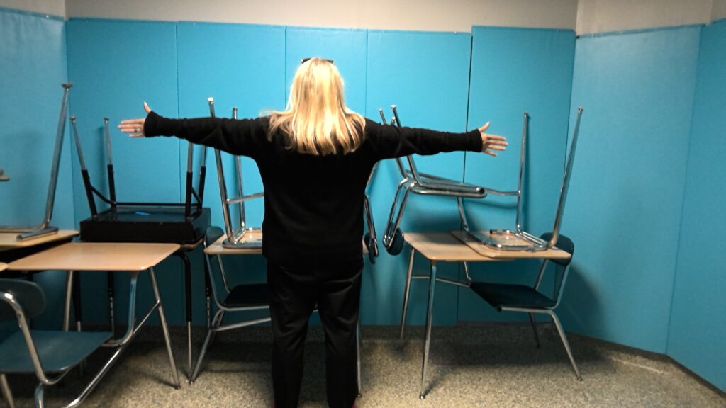 An adult stands in a room with padded walls and uncarpeted floors. In the room are multiple student desks stacked 2-high.