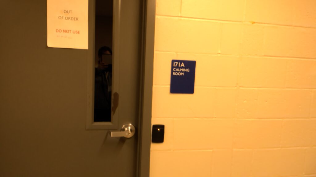 A metal door with narrow observation window. A plaque next to the door reads "171A Calming Room." A sheet of paper attached to the door reads "OUT OF ORDER DO NOT USE"
