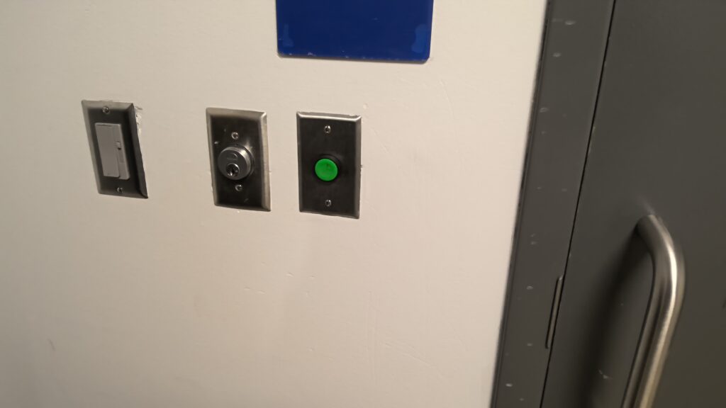 Photograph showing a section of a white wall with three control switches: a rectangular light switch, a round key-operated switch, and a green push button. A gray door with a metal handle is partially visible on the right side, indicating an access or control point.
