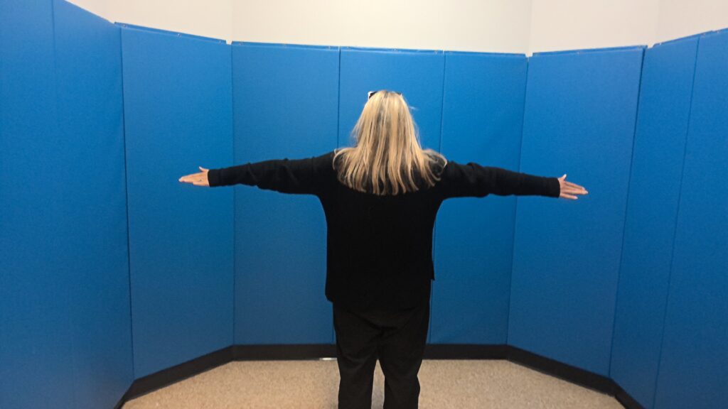 An adult stands in the center of a room with blue padded walls and uncarpeted floors.