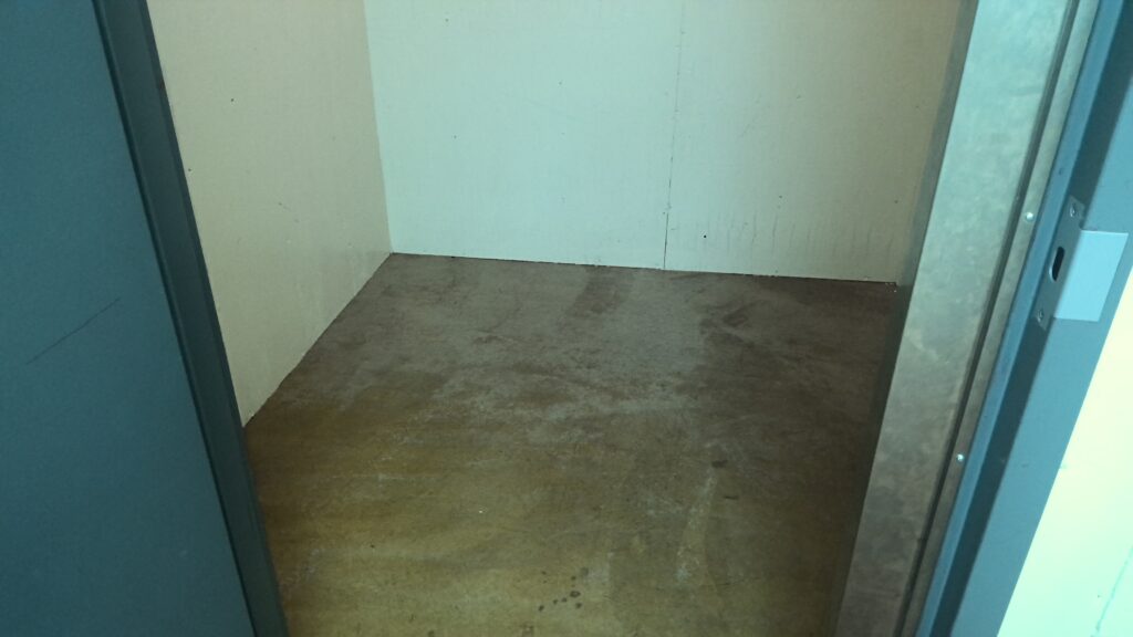 Small room with white paneled walls and concrete floor viewed through a metal doorway.