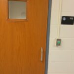 Wood door with small square observation window. Green button on wall next to door.