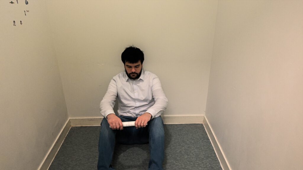 An adult sits on the floor with their back against the wall in a small room with white walls and gray flooring. The wall is only about 3 times the width of the adult's body.