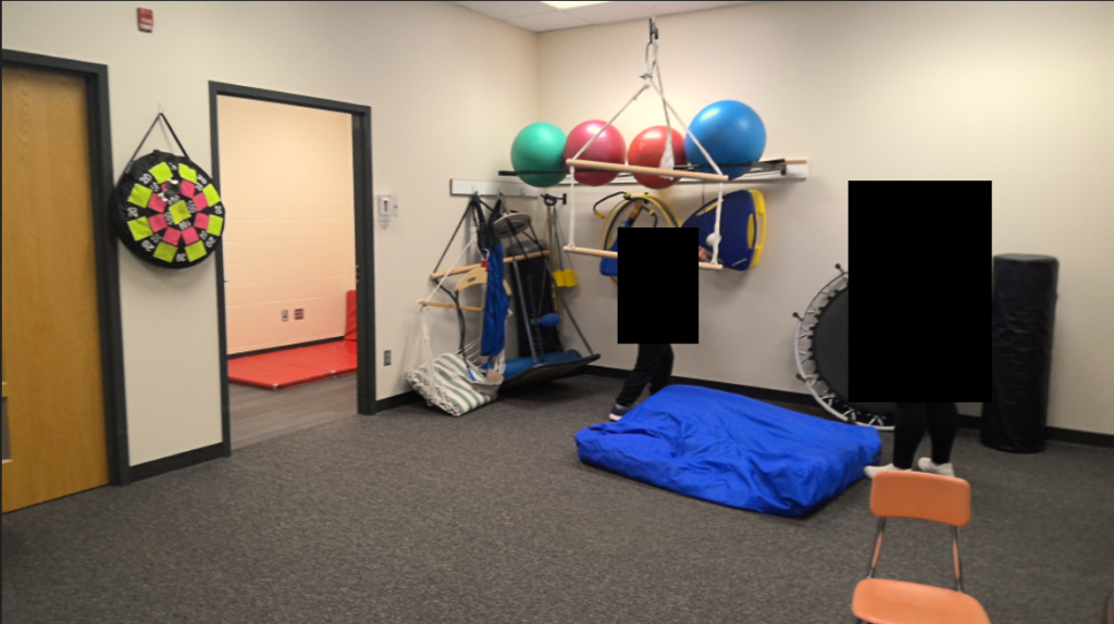 An adult and child standing in a room with miscellaneous occupational therapy equipment.