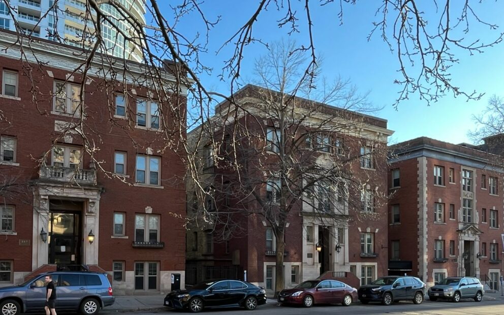 Street view of three red brick, 3-story apartment buildings stand side-by-side with a high-rise building in the background on a sunny spring day