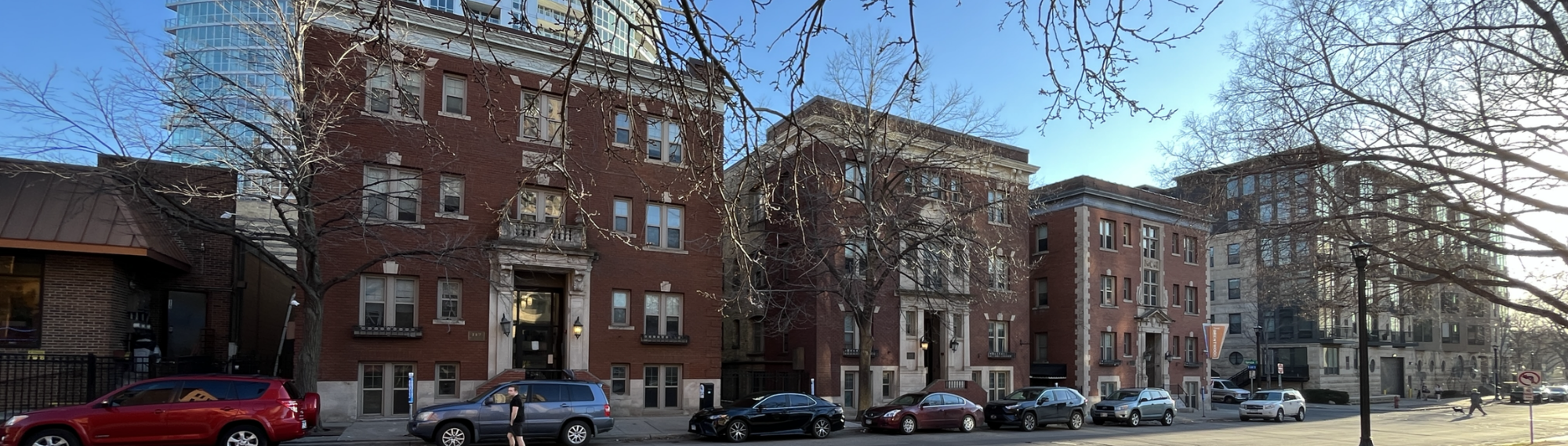Street view of three red brick, 3-story apartment buildings stand side-by-side with a high-rise building in the background on a sunny spring day