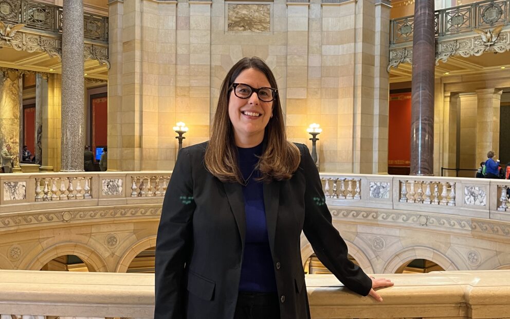 Woman with long brown hair, glasses, in professional attired, black blazer, pants, stands with the backdrop of the warmly lit Capitol rotunda behind her.