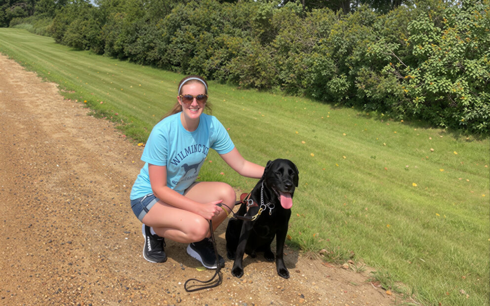 Teenager in sunglasses wearing blue shirt and denim shorts kneels next to dog in sunny outdoor setting on dirt trail next to grass
