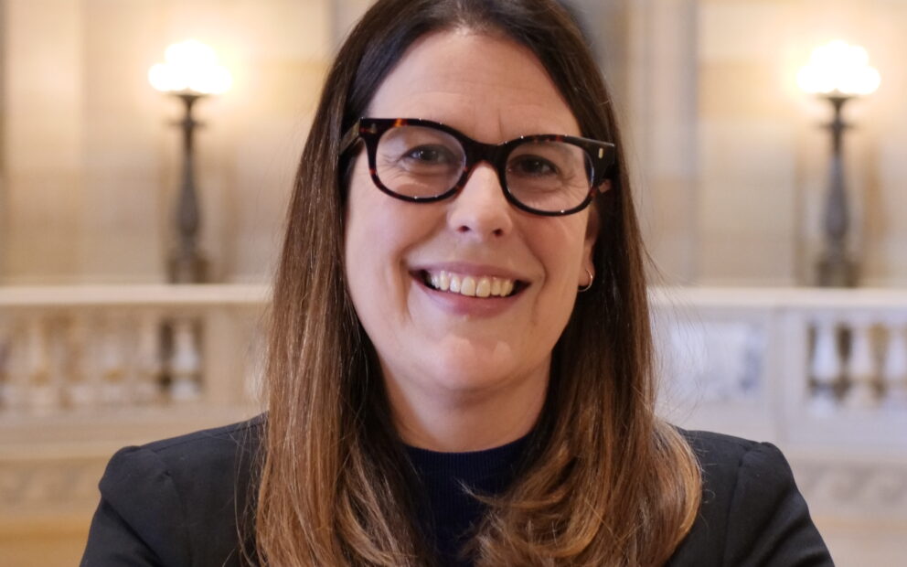 Woman with brown, longn hair, wearing dark-rimmed glasses and ams crossed smiles with the background of the MN Capitol Rotunda behind her