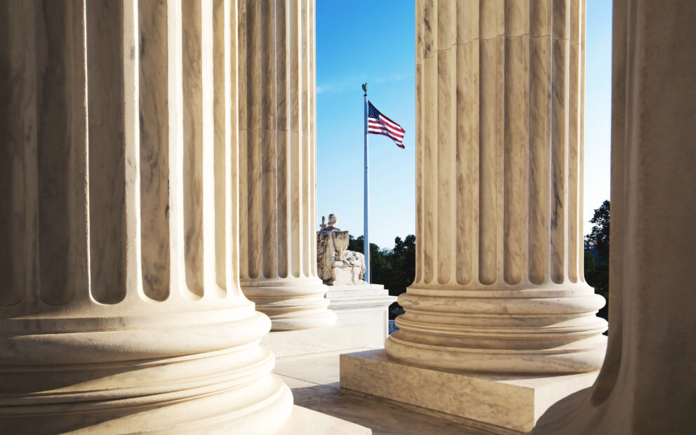 Between pillars of the US Supreme Court building is view of US Flag in the distance beneath a blue sky.
