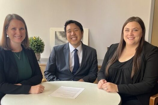 Three individuals in suits/blazers sit at a small conference table, smiling