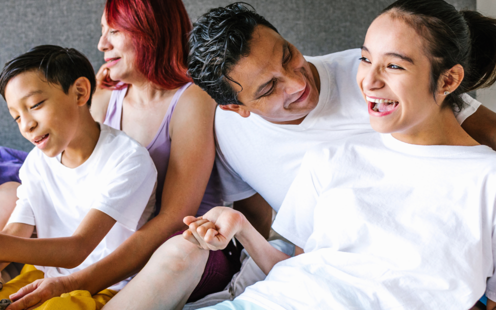 Family sitting in living area, mother holds school-age son; father peers over shoulder of daughter with musculoskeletal condition. All smiling.