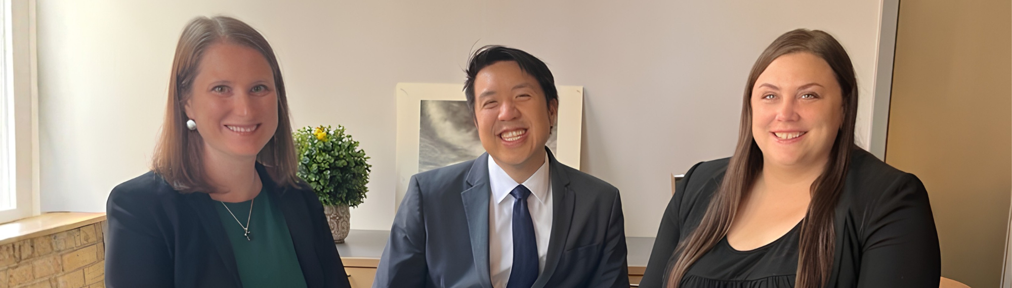 Three individuals in suits/blazers sit at a small conference table, smiling