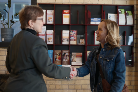Woman enters lobby door extends hand to another who is also extending a hand as they meet.