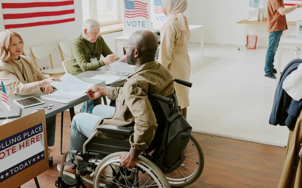 Election workers seated on one side of table face an individual in wheelchair on opposite side of a voter check-in table