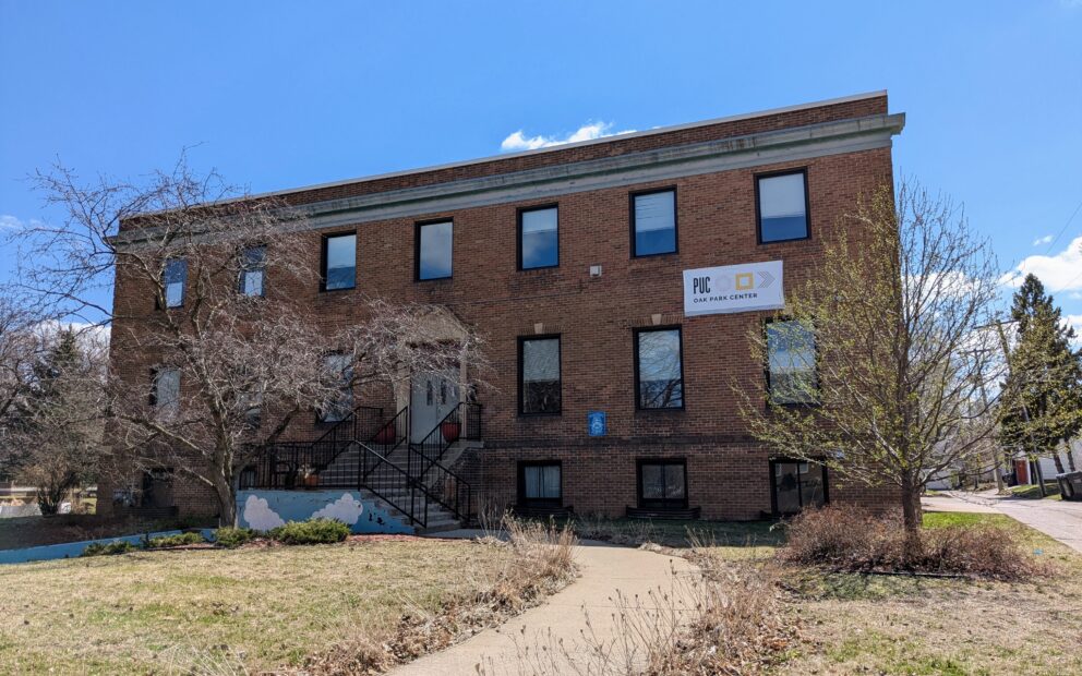 Wide shot of two-story, red brick building, lawn in front with sidewalk to building; set against blue sky in background